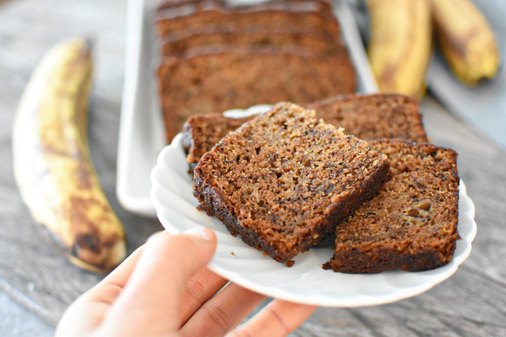 hand holding a slice of banana bread on a plate with banana bread in the background